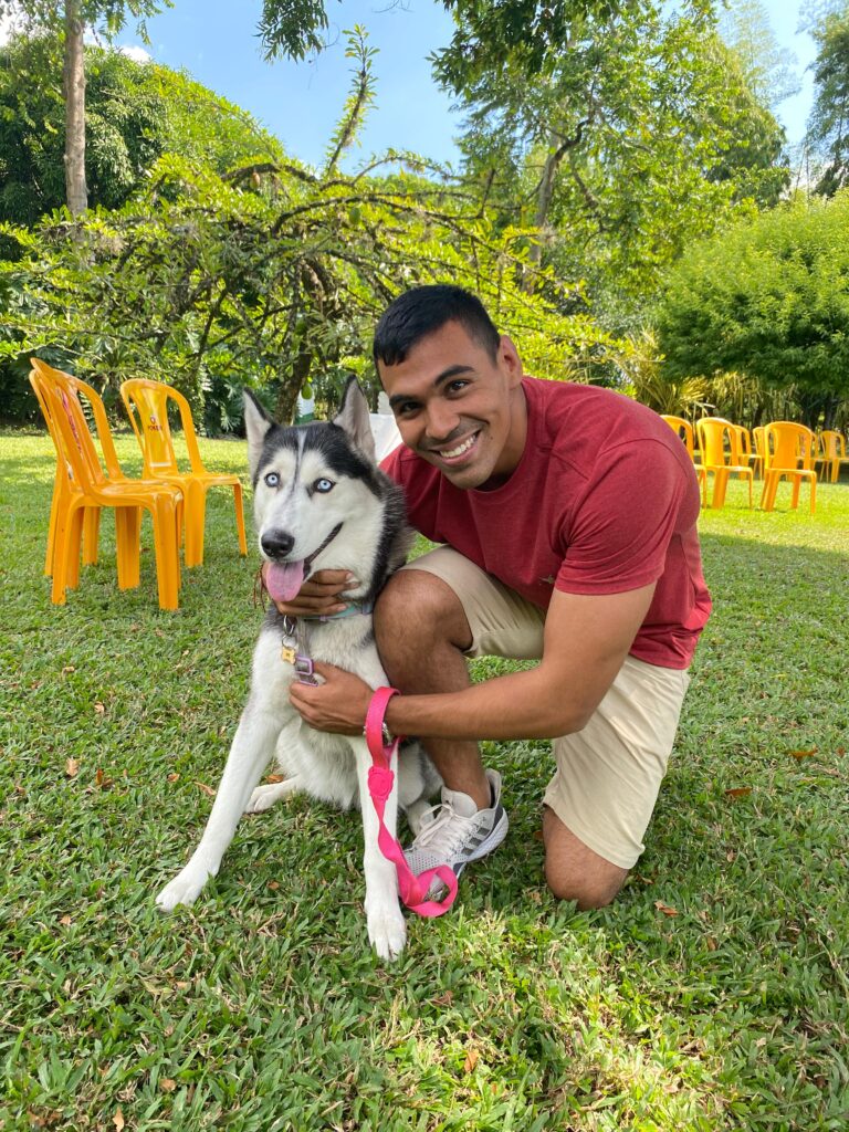 Un hombre jóven mira a la cámara abrazando a su perra Husky siberiano en un entorno rural, rodeado de árboles con un cielo despejado y sillas de color amarillo.