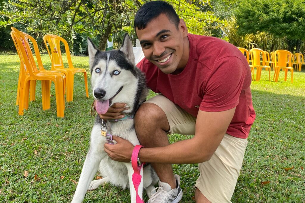 Un hombre jóven, Juan Sebastián Bernate, mira a la cámara abrazando a su perra Husky siberiano en un entorno rural, rodeado de árboles con un cielo despejado y sillas de color amarillo.