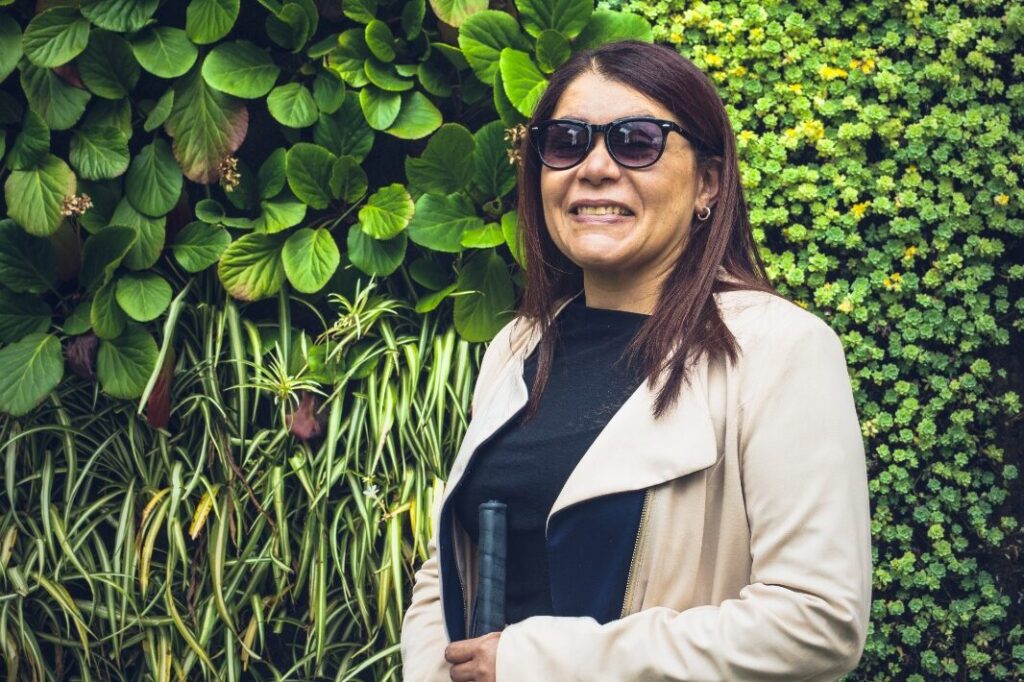 Yecenia Fernández, está parada sosteniendo su bastón, que es de color verde neón y detrás de ella hay una pared forrada en un jardín vertical con distintos tonos de verde. Lleva puesta una gabardina color beich, blusa negra, gafas de sol y sonríe mirando a la cámara