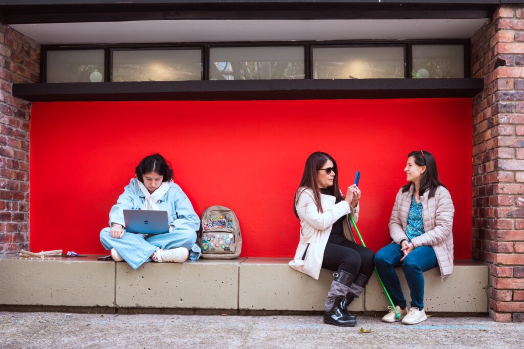 En la parte izquierda de la imágen, sentadas sobre un muro sobresaliente de cemento, Yecenia Fernández, una mujer ciega, enfoca la cámara de su celular hacia una mujer jóven que la mira y le sonríe, es la entrevistadora. Se ve su bastón verde neón y tras de ellas una pared de color rojo encendido. Al lado derecho de la imágen una mujer, sentada sobre el mismo muro hay una mujer jóven, estudiante, concentrada en la pantalla de su computador.