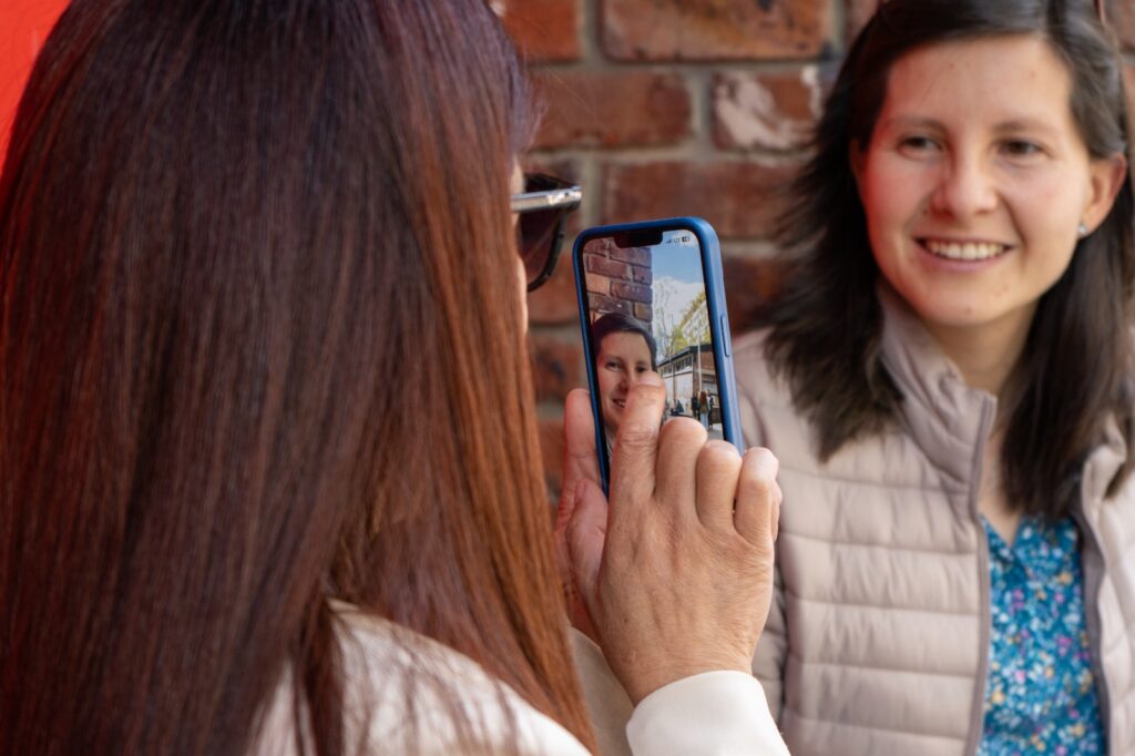 A la izquierda, Yecenia Fernández, una mujer ciega que da la espalda a la cámara, le toma una foto con su celular a Juliana Mancera, la periodista que la está entrevistando y que se ubica al lado derecho de la imágen. En la pantalla del celular de Yecenia se amplía el colorido fondo que rodea a Juliana, un cielo despejado y soleado, edificios en diferentes planos rodeados de árboles.