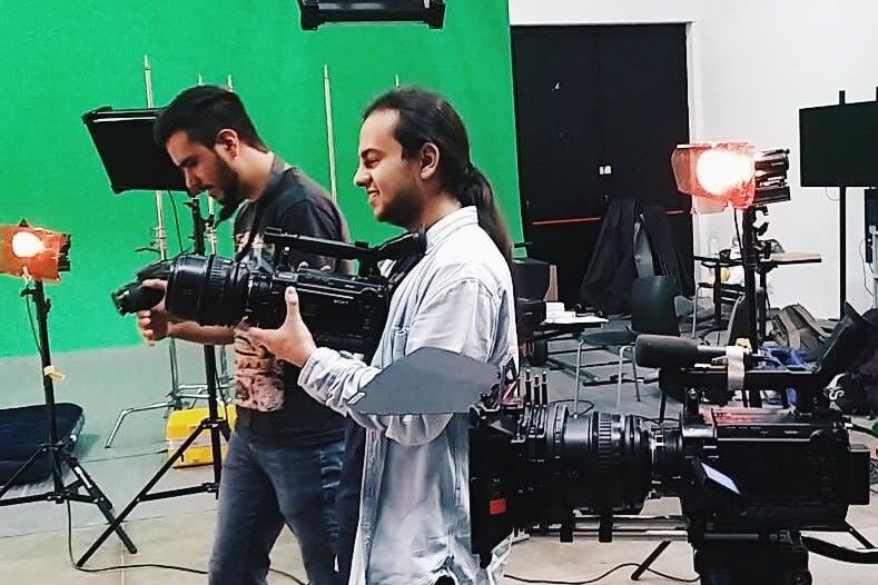 Juan Camilo Montaña, en un estudio de grabación con pantalla verde al fondo, sostiene en sus manos una cámara cinematográfica mientras sonríe