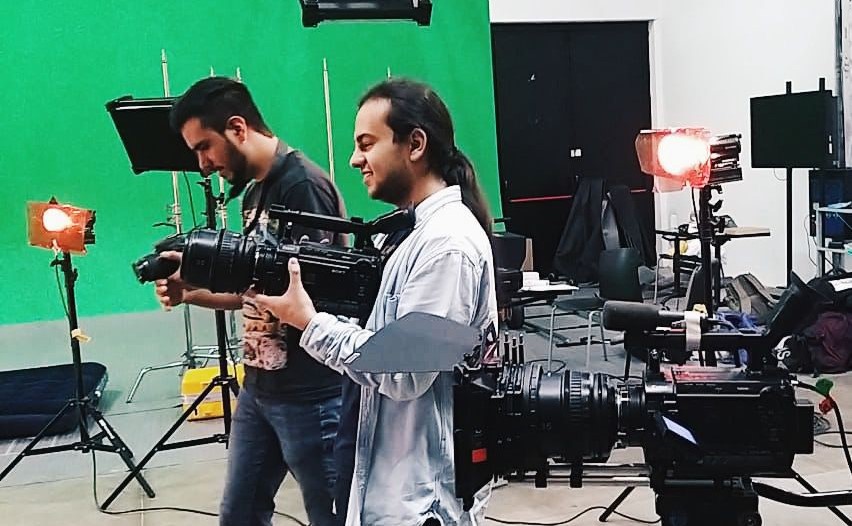 Juan Camilo Montaña, en un estudio de grabación con pantalla verde al fondo, sostiene en sus manos una cámara cinematográfica mientras sonríe